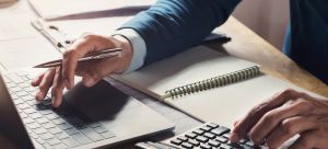 Schreibtisch mit Laptop, Notizbuch und Taschenrechner. Eine männliche Person hält in er rechten Hand einen Kugelschreiber und tippt auf dem Laptop. Mit der linken Hand tippt er auf dem Taschenrechner.